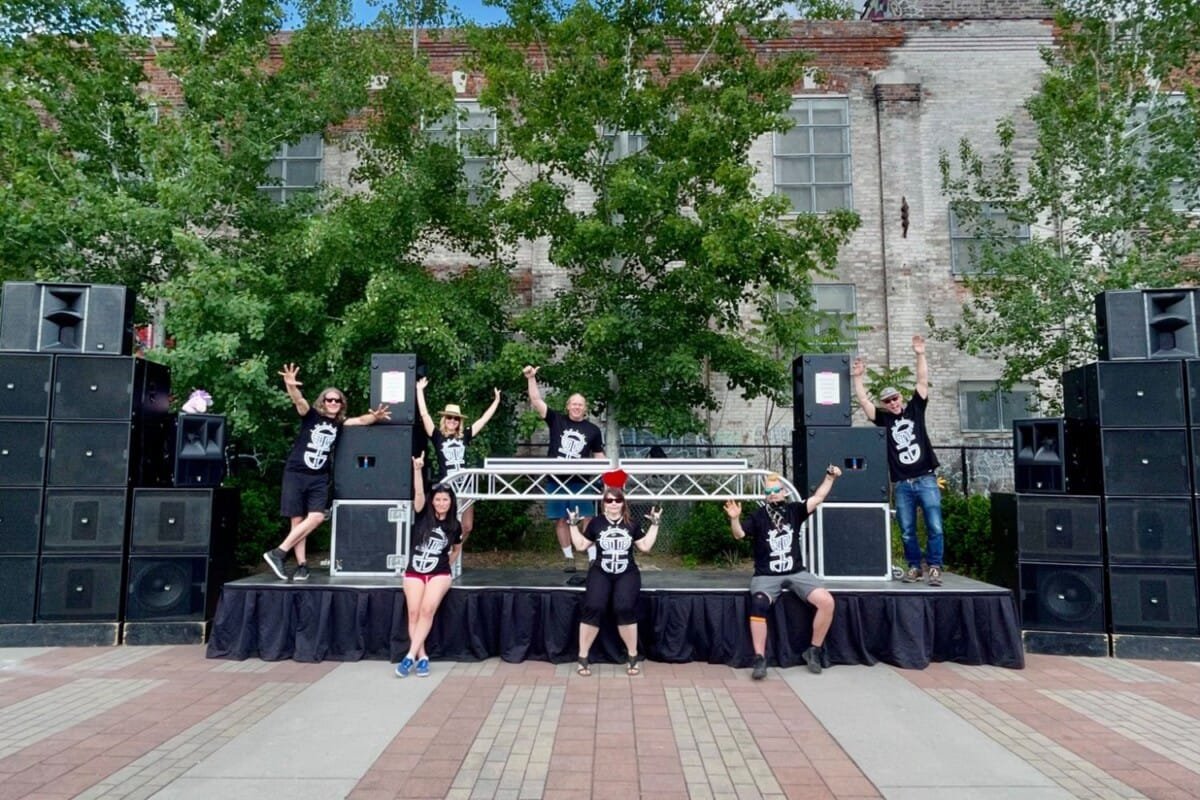 Seven people with arms in the air in front of a an outdoor stage surrounded by sound system rental speakers