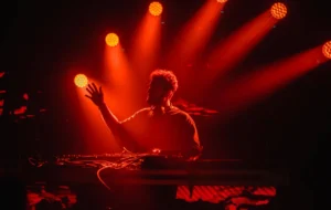 A curly haired DJ lit by 6 red spotlights and has one hand in the air lifted up