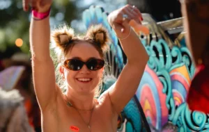 A woman with furry cat ears, sunglasses, and hands in the air. A SPICY orange sticker on her upper breast
