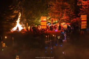 A crowd at night next to a bonfire and barge. Flags at side.