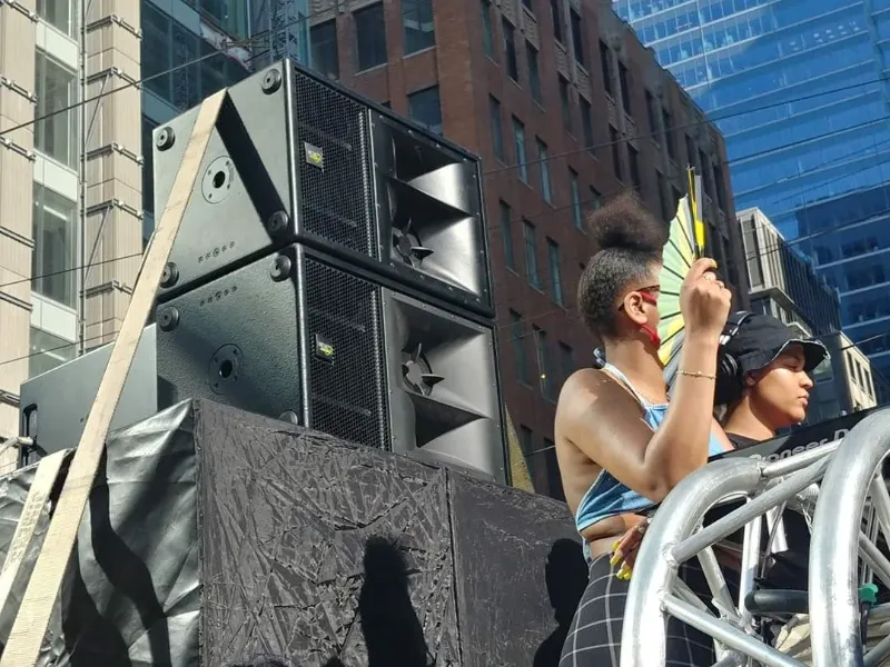 Two speakers on two sub-woofers mounted on a truck with a woman with naturally curly hair and holding a fan, and a DJ wearing a hat performing beside them.