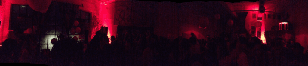 A panoram of a crowded red lit room of dancers