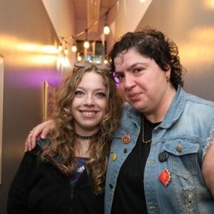 Two people smiling in a corridor with warm hanging lights. One wears glasses on their head and a black jacket; the other has short curly hair, a denim waistcoat with badges, and their arm around the first person.