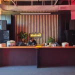 A DJ stands behind a wooden counter with audio equipment in a modern room lit by red lights, large speakers on both sides, and a neon sign on a wooden panel backdrop. Another person adjusts equipment on the right.