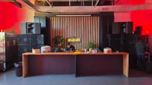 A DJ stands behind a wooden counter with audio equipment in a modern room lit by red lights, large speakers on both sides, and a neon sign on a wooden panel backdrop. Another person adjusts equipment on the right.