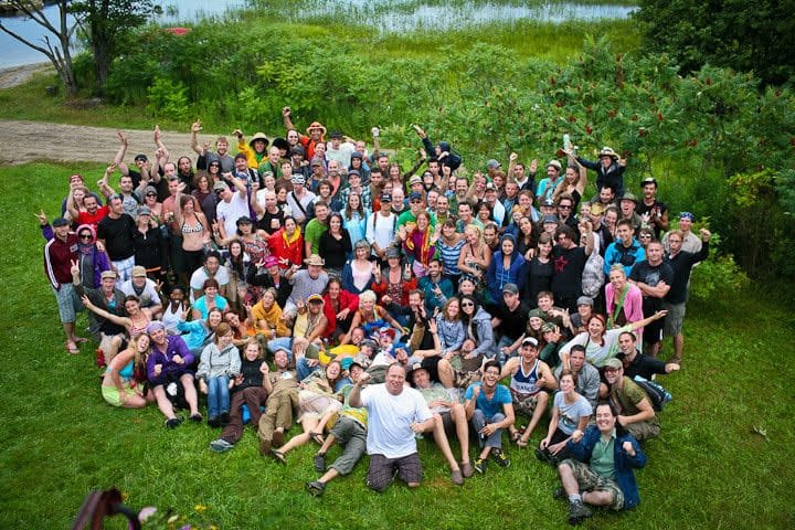 A large, diverse group of people gathered outdoors on green grass, smiling and posing together for a photo near trees and a body of water in the background. Many have their arms raised and seem joyful.