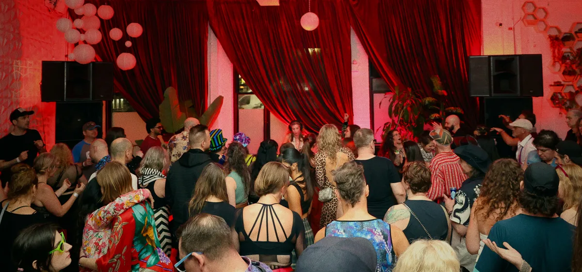 A crowd in colourful clothing gathers in a dimly lit room with red curtains, balloons, and plants for a lively Sunday afternoon social, all facing a performer on stage. The festive atmosphere buzzes with excitement.