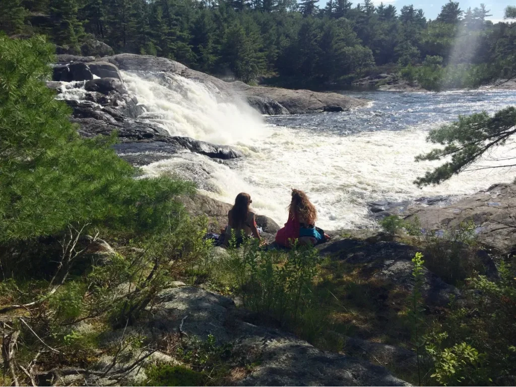 Two people sit on a rocky, grassy area beside a rushing river and waterfall, surrounded by boreal pine trees and natural greenery under a bright, sunny sky.