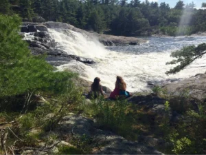 Two people sit on a rocky, grassy area beside a rushing river and waterfall, surrounded by boreal pine trees and natural greenery under a bright, sunny sky.