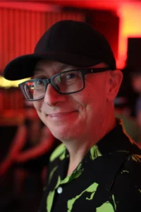 Man in black baseball cap and glasses smiles in a neon-lit room.