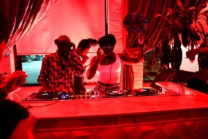 DJs in red lighting with turntables and speakers at a party