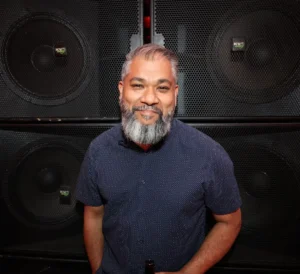 DJ with a beard smiles in front of large speakers at a music event.