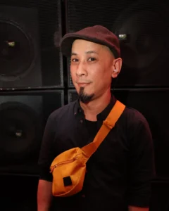 DJ with goatee, flat cap, and orange fanny pack in front of speakers