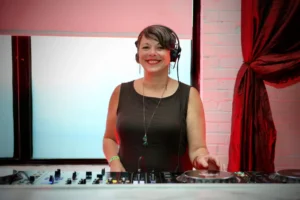 Female DJ with headphones smiles behind a DJ setup at an event.