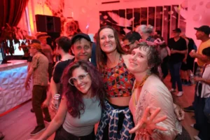 Four smiling people at a lively party with colorful decor, possibly a music venue.