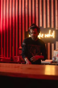 DJ wearing headphones plays music at a sushi bar with neon "sushi" sign.