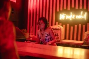 Woman in cow print bikini and glasses behind bar with neon "let's make sushi" sign