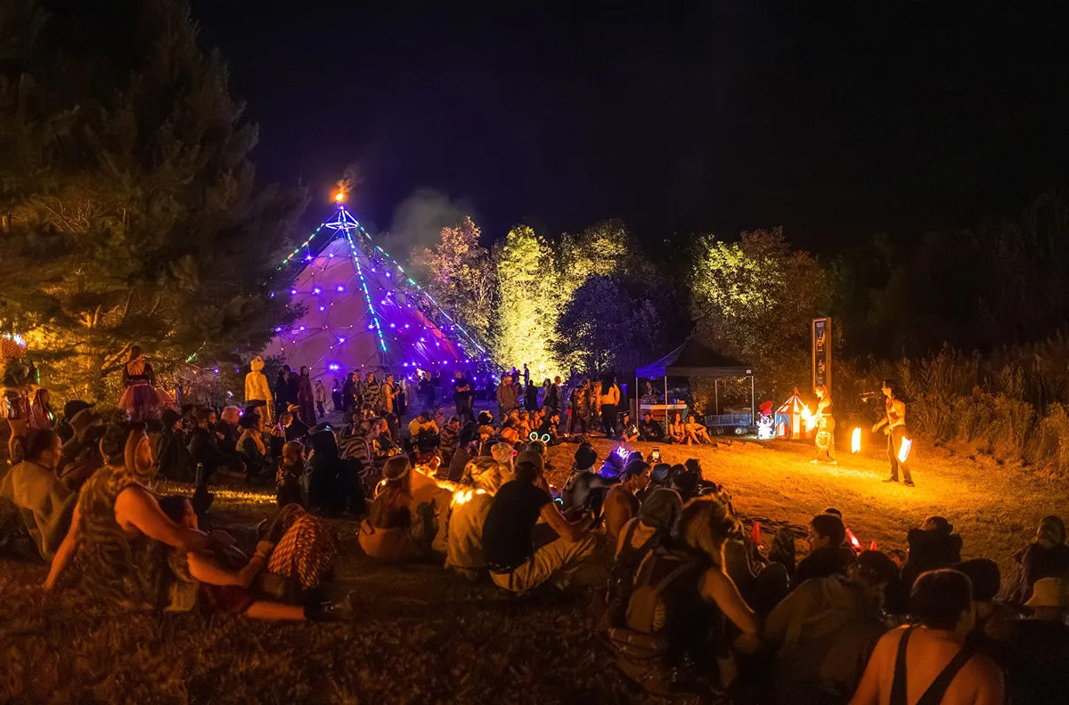 Festival crowd at night around a illuminated teepee with fire performers