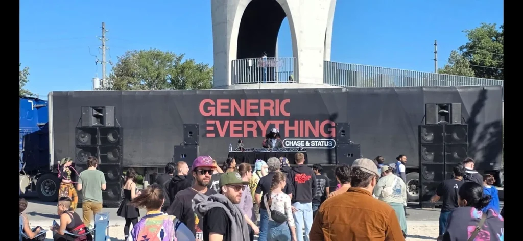 DJ performing on stage with "GENERIC EVERYTHING" banner, speakers, and early crowd